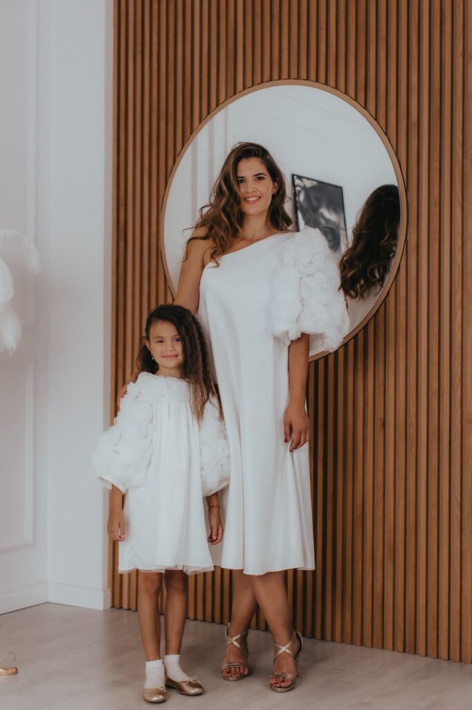 Postoje trenuci koji su tihi, ali nezaboravni — intimna venčanja, krštenja i posebni porodični događaji u kojima elegancija dolazi iz jednostavnosti. Svečane haljine za mamu i ćerku „Vjeruj“ u beloj boji stvorene su upravo za takve prilike, kada želite da izgledate skladno, nežno i posebno. Haljina „Vjeruj“ za žene odiše čistom elegancijom i sofisticiranim dizajnom. Bela boja naglašava njenu bezvremensku ljepotu, dok asimetričan rukav ukrašen ručno rađenim ružama od tila daje poseban karakter i čini ovu svečanu haljinu jedinstvenom. Kroj prati liniju tela i ističe ženstvenu siluetu na suptilan način. Svečana haljina za devojčice „Vjeruj“ donosi isti osjećaj elegancije u nježnoj, prilagođenoj formi. Lagani materijali omogućavaju udobnost i slobodu kretanja, dok detalji u vidu ruža od tila unose razigranost i posebnu čar, čineći svaki trenutak još ljepšim. Ovaj set haljina za mamu i ćerku donosi više od usklađenog izgleda — donosi osjećaj povezanosti, nježnosti i zajedničkih trenutaka koji ostaju zabeleženi kroz fotografije i uspomene. To su trenuci u kojima ste zajedno, elegantne i usklađene, u savršenom balansu jednostavnosti i posebnosti. Izrađene sa detaljima od tila i ručno rađenim elementima, ove svečane haljine za mamu i ćerku spajaju minimalizam, eleganciju i emociju u jedan skladan komplet. Ključne karakteristike: Usklađen set haljina za mamu i ćerku Bela boja kao simbol elegancije i bezvremenosti Asimetričan rukav sa ručno rađenim ružama kao upečatljiv detalj Lagani materijali koji omogućavaju udobnost i pokret Minimalistički i sofisticiran dizajn Idealno za venčanja, krštenja i posebne porodične trenutke