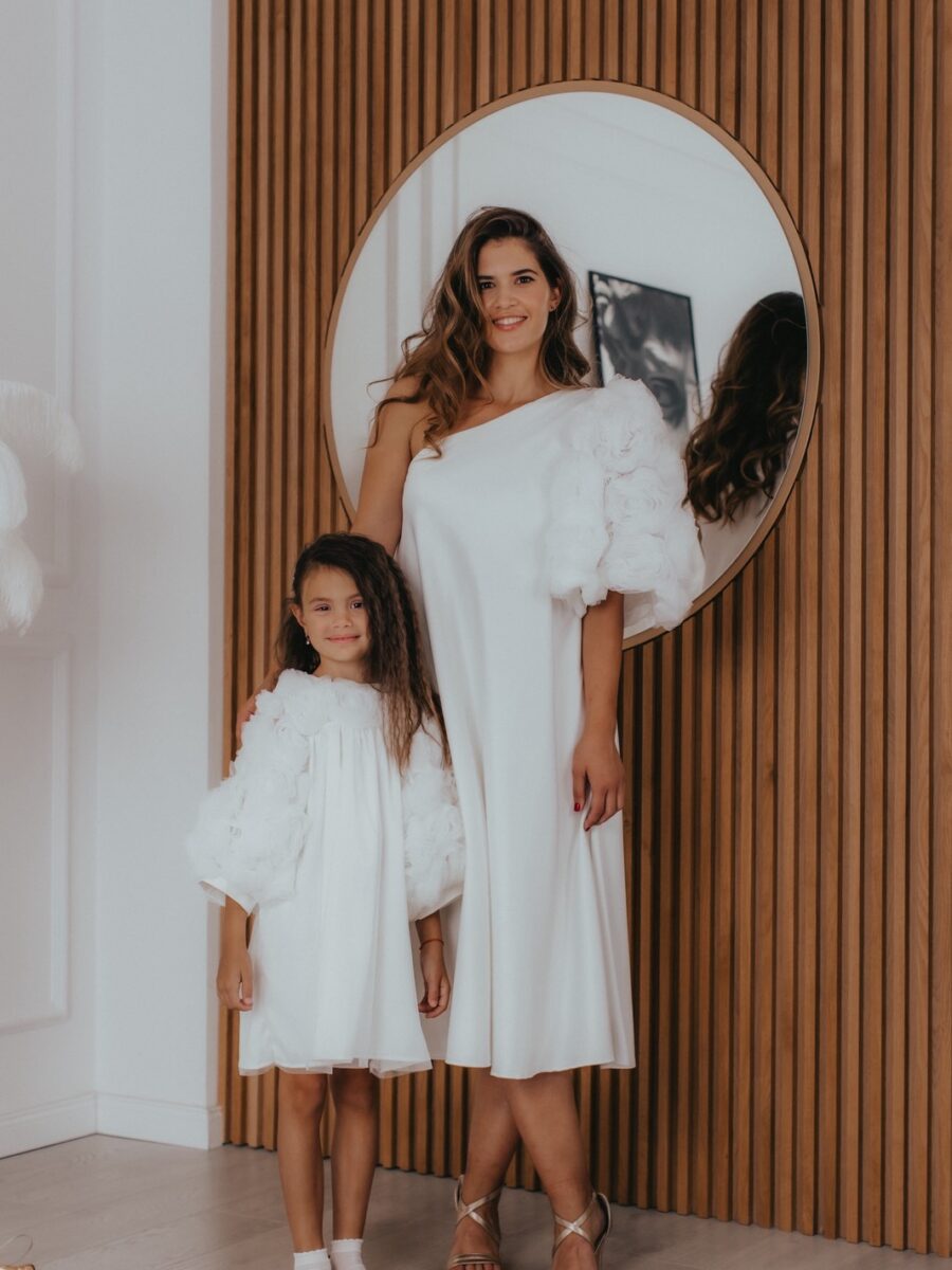 Postoje trenuci koji su tihi, ali nezaboravni — intimna venčanja, krštenja i posebni porodični događaji u kojima elegancija dolazi iz jednostavnosti. Svečane haljine za mamu i ćerku „Vjeruj“ u beloj boji stvorene su upravo za takve prilike, kada želite da izgledate skladno, nežno i posebno. Haljina „Vjeruj“ za žene odiše čistom elegancijom i sofisticiranim dizajnom. Bela boja naglašava njenu bezvremensku ljepotu, dok asimetričan rukav ukrašen ručno rađenim ružama od tila daje poseban karakter i čini ovu svečanu haljinu jedinstvenom. Kroj prati liniju tela i ističe ženstvenu siluetu na suptilan način. Svečana haljina za devojčice „Vjeruj“ donosi isti osjećaj elegancije u nježnoj, prilagođenoj formi. Lagani materijali omogućavaju udobnost i slobodu kretanja, dok detalji u vidu ruža od tila unose razigranost i posebnu čar, čineći svaki trenutak još ljepšim. Ovaj set haljina za mamu i ćerku donosi više od usklađenog izgleda — donosi osjećaj povezanosti, nježnosti i zajedničkih trenutaka koji ostaju zabeleženi kroz fotografije i uspomene. To su trenuci u kojima ste zajedno, elegantne i usklađene, u savršenom balansu jednostavnosti i posebnosti. Izrađene sa detaljima od tila i ručno rađenim elementima, ove svečane haljine za mamu i ćerku spajaju minimalizam, eleganciju i emociju u jedan skladan komplet. Ključne karakteristike: Usklađen set haljina za mamu i ćerku Bela boja kao simbol elegancije i bezvremenosti Asimetričan rukav sa ručno rađenim ružama kao upečatljiv detalj Lagani materijali koji omogućavaju udobnost i pokret Minimalistički i sofisticiran dizajn Idealno za venčanja, krštenja i posebne porodične trenutke